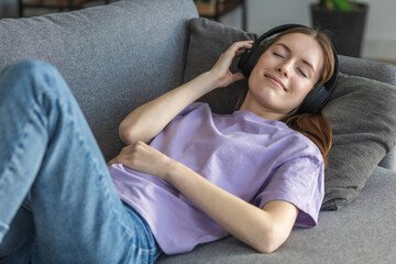 Obraz premium Relax at home, positive thinking. Happy young woman lying at home on the couch with headphones listening to her favorite music and thinking about something good, smiling