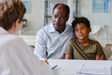 African dad with his little son sitting at table and listening to prescription of doctor at office