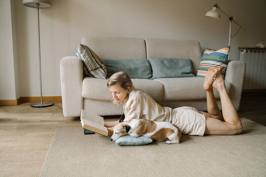Attractive blonde woman enjoying leisure time reading novel home with small friend dog Jack Russell terrier sleeping. Relaxed moments chilling at home with your pet. Slow life atmosphere vibe