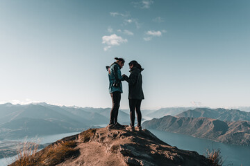 caucasian boy with long collected hair next to brunette caucasian girl on top of a mountain talking in natural environment near big blue lake and rocky mountains, roys peak, new zealand - Powered by Adobe