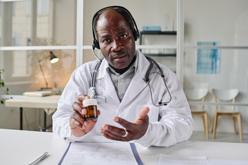 Portrait of African mature pediatrician in headphones giving online consultation about pills while sitting at table at office