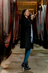 Woman in the wine cellar with barrels in background drinking and tasting wine.