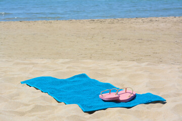 Soft light blue beach towel and pink flip flops on sand near sea. Space for text