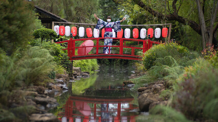 Japanese women with masks and beautiful dresses casual Yukata Kimono and red umbrellas wear traditional clothes and stand on a red wooden bridge with red and white floral lanterns in garden.