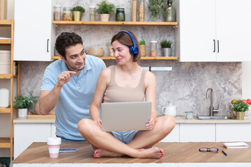 Young married couple amusing in the kitchen. Attractive wife sitting on the table and working on laptop and her husband cleaning the kitchen