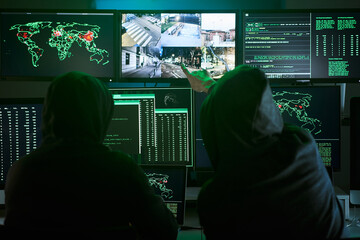Back view of unrecognizable Criminal team Hacker pointing security cameras on screen monitor using computer for organizing massive data breach attack on government or big company servers. High quality