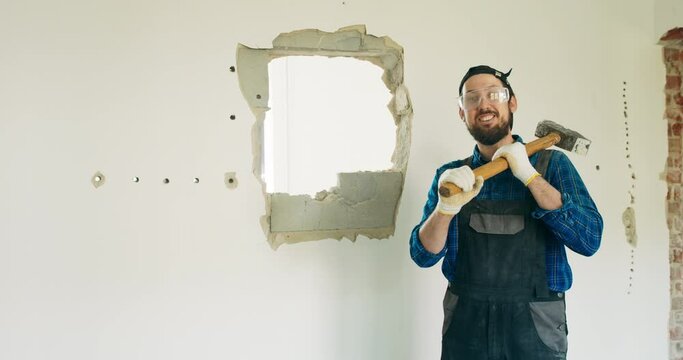 A man working on a construction site wearing a work suit with a visor and safety goggles in his hands holds a hammer with which he demolishes a wall smiles at the camera.