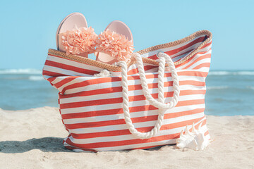 Stylish striped bag with slippers and seashell on sandy beach near sea