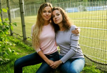 Two women are sitting on the grass in the park next to the football field.