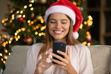 Happy young beautiful woman in festive red hat using cellphone, reading message with New Year greetings communicating in social network, sitting on sofa near decorated Christmas tree at home.