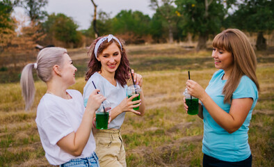 Fototapeta premium Three young women in fashionable clothes, out of town in nature, in summer and drinking refreshing non-alcoholic cocktails from disposable plastic cups. Female friendship. Holidays, pleasant leisure.