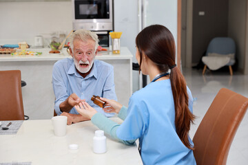 Happy caregiver sitting and helping senior caucasian man taking medicine after breakfast in living room at retirement house. Asian smiling nurse taking good help care and support elder patient at home