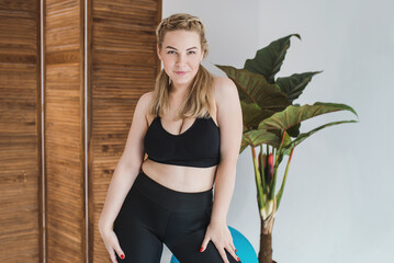 A plus size woman in a black sports top and leggings. Chubby blonde is engaged in fitness. Curvy girl posing  in a bright room 