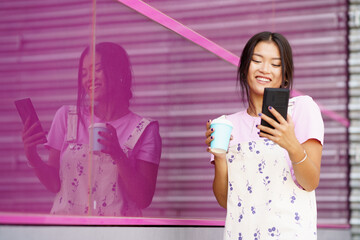 Asian girl near a modern pink building, drinking coffee in a take-away cup, checking her smartphone