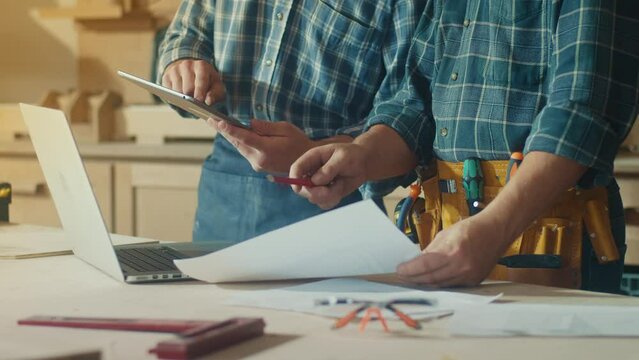 Small Business Owners of a Furniture Workshop Using Tablet, Notebook and Discussing the Design of a New Wooden Product. Hands of Carpenter and Young Male Apprentice Working in Studio.