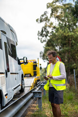 Caucasian man waiting tow truck motor home on the highway with a reflective vest and checking his...
