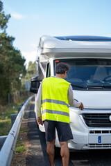 Caucasian man waiting a mechanic to come fix his broken down motor home on the highway with a reflective vest and checking the vehicle