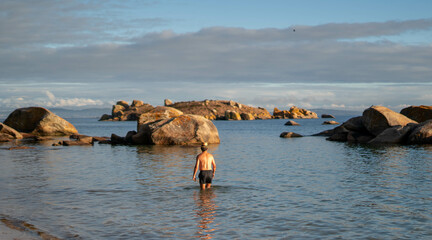 Caucasian man entering at water with a hat and blue swimsuit with rocks in the background