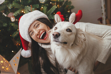 Merry Christmas! Happy woman in santa hat hugging funny white danish spitz dog in festive room....