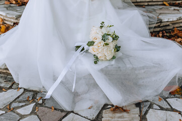 A beautiful wedding bouquet of white roses with a ribbon lies on the bride's long dress. Close-up photography, portrait.