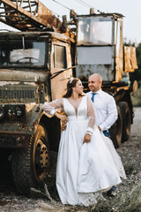 A stylish adult groom and a beautiful bride in a white dress stand embracing against the backdrop of an old, rusty truck car in ruins in Ukraine. wedding photography.