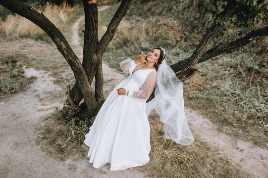 A beautiful, young, smiling bride in a white dress stands in nature in a park near a tree. Wedding photography, portrait.