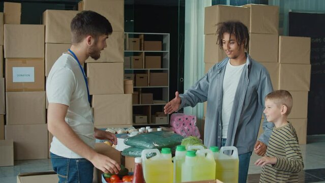 Friendly Volunteer Giving Donation Box With Food to Man and His Son at Distribution or Refugee Assistance Center. The Concept of Social Aid. Internally Displaced Persons