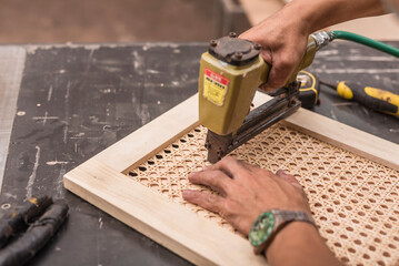 A man fastens a rattan solihiya weave screen onto a cabinet panel with a pneumatic staple gun. At a...