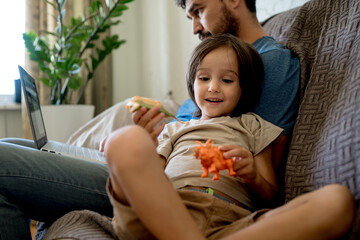 The father works at home on a laptop and the child requires attention, wants dad to rub toys with him.