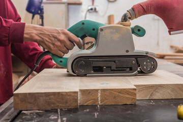 Using a hand-held sanding machine or belt sander to level the surface of a glued wood for a cabinet, table or other furniture at a workshop.