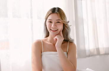 Obraz na płótnie Canvas Happy beautiful Asian girl with her finger raised to her cheek and looking at camera while sitting in the morning with sunbeam dawn on her face in bedroom. Pretty Korean woman clean fresh skin concept