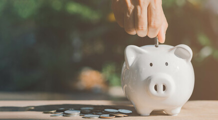 Person hand putting coins into white piggy bank in sun shine morning with copy space, coins are on wooden table. Saving money, investment, fund, loan, education, healthy, insurance, banking concept.