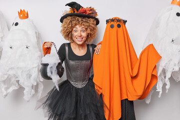 Surprised cheerful woman witch poses cauldron smiles gladfully wears black hat and dress stands near spooky ghost enjoys party on Halloween night poses around spooky creatures. 31st of October