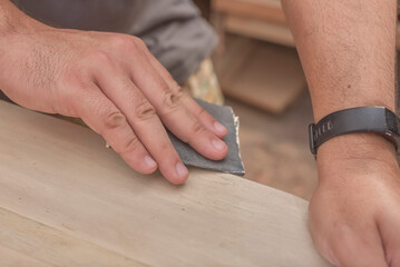 A man uses a piece of sandpaper to smooth out the edge of a circular table. Hand sanding a wood surface. A DIY project at a personal workshop.