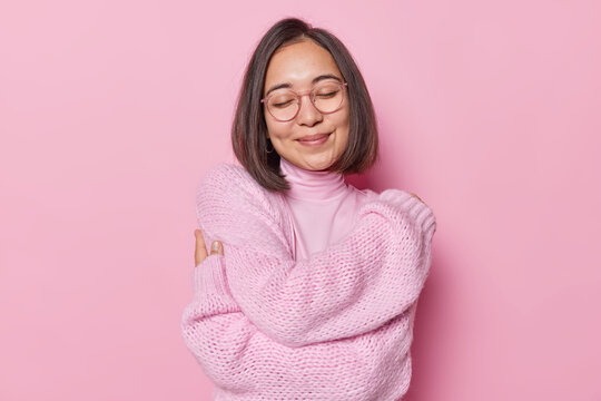 Tender Asian woman with dark hair embraces herself closes eyes and smiles lovely wears transparent glasses comfortable jumper feels cozyness isolated over pink background recalls romantic date
