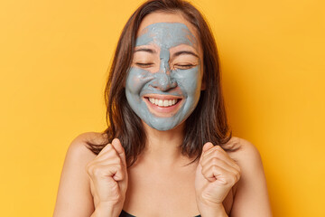 Horizontal shot of positive dark haired woman clenches fists keeps eyes closed smiles gently applies facial clay mask for skin care has well cared body stands bare shoulders isolated over yellow wall
