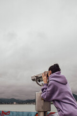Obraz premium a girl watching through stationary binoculars on the pier for passing ships and wildlife on the seashore