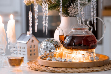 A bouquet of fir branches with icicles, candles and a teapot with tea on a wooden table in the home interior of a white Scandinavian-style kitchen. Cozy concept of festive drinks.