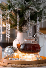 A bouquet of fir branches with icicles, candles and a teapot with tea on a wooden table in the home interior of a white Scandinavian-style kitchen. Cozy concept of festive drinks.