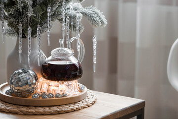 A bouquet of fir branches with icicles, candles and a teapot with tea on a wooden table in the home interior of the living room. Cozy concept of festive drinks.