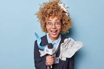 Positive surprised curly haired female journalist or TV presenter speaks into microphone wears round eyeglasses and formal clothes exclaims loudly isolated over blue studio background. News media