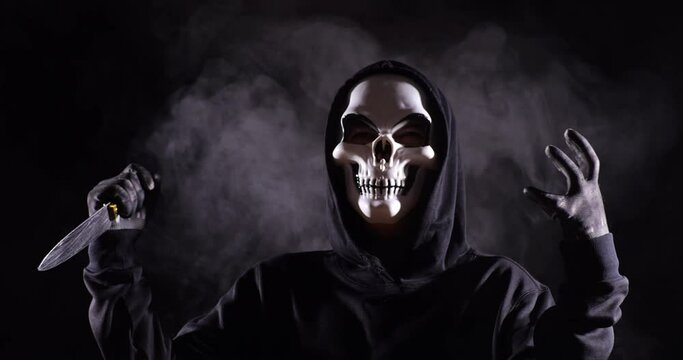 Close Up Of Crazy Man In The Hooded Sweatshirt Wearing Halloween Mask Holding A Knife And Laughing On The Black Background With Smoke
