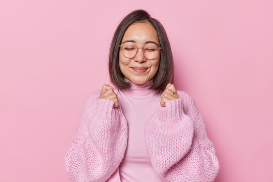 Portrait of Asian woman with dark straight hair keeps eyes closed smiles gently clenches fists awaits for something good happen wears transparent eyeglasses and jumper isolated over pink background