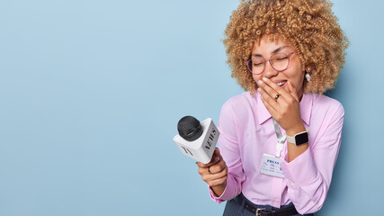 Overjoyed curly haired female reporter covers mouth laughs gladfully holds microphone participates...