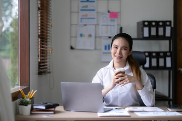 Beautiful young Asian businesswoman smiling holding a coffee mug and laptop working at the office.