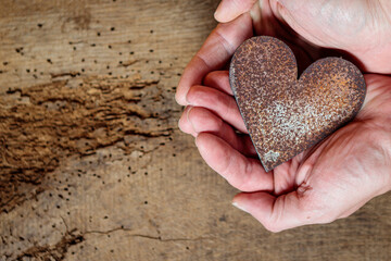 Zwei Hände halten ein Herz aus rostigem Stahlblech vor rustikalem Eichenholzbrett.