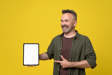 bristle and mustached mature man dressed in green shirt happy smiling pointing with thumb up and finger to tablet pc gadget with white mock-up screen holding in hand on yellow background