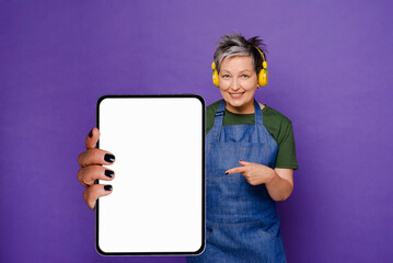 Great Mobile Offer. Smilling grey haired Lady Pointing Finger at big tablet pc in hand isolated over violet studio background. Happy woman showing touchpad tablet pc, with white screen copy space area
