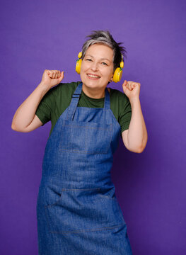 Portrait Of Attractive Middle Aged Woman In Blue Jeans Apron Dancing With Music In Her Yellow Headphones Isolated. Mature Happy Housewife Loves Dancing Music Over Violet Background