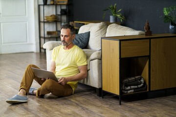 Middle-aged Caucasian man relaxing at home sitting on flow lean on sofa couch with laptop, e-learning, watching seminars, videos, movies online remotely. Freelance work concept